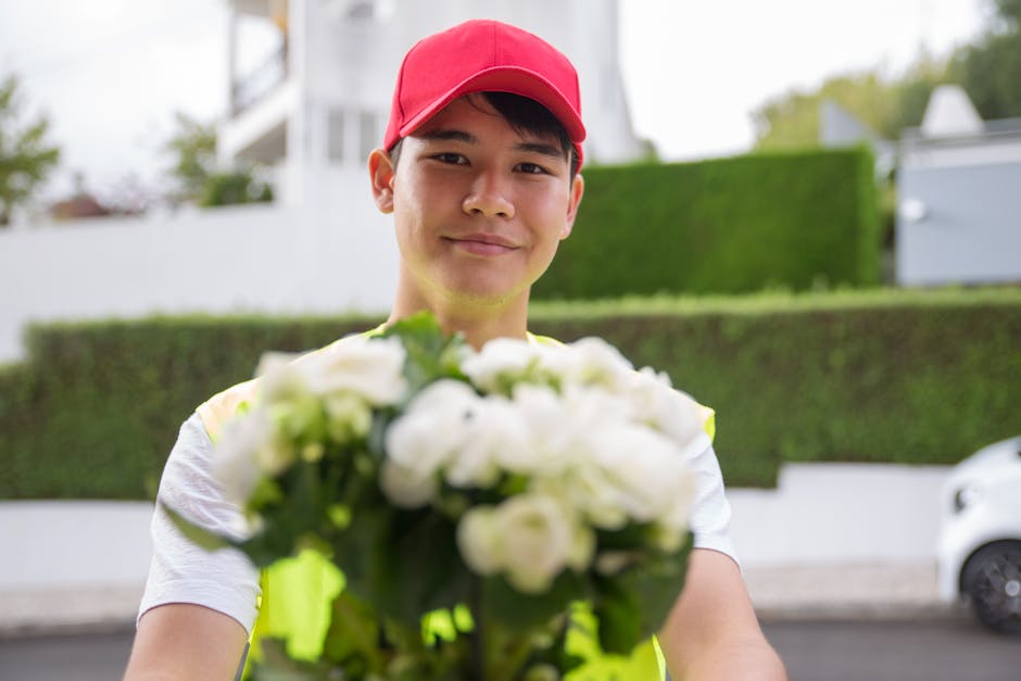A young delivery person with light skin, wearing a red cap and a yellow high-visibility vest, is smiling while holding a bouquet of fresh white roses and greenery. The bouquet is arranged neatly with 