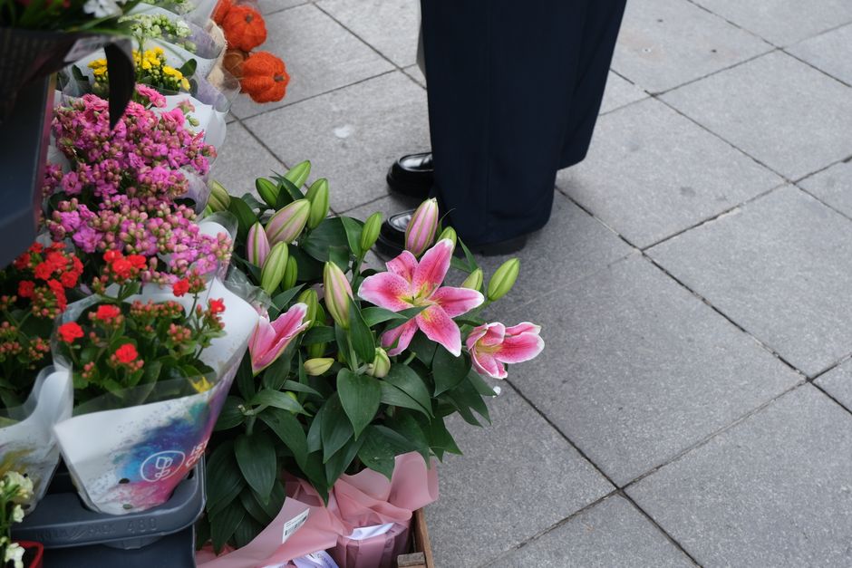 A display of freshly cut flowers on a flower stall outside a shop, including pink and white lilies with open blossoms and closed buds, surrounded by green foliage, set against wrapping paper and ribbo