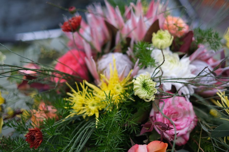A vibrant floral arrangement featuring a variety of fresh flowers, including pink roses, yellow spider chrysanthemums, white ranunculus, and green foliage, positioned together in a natural, loose bouq