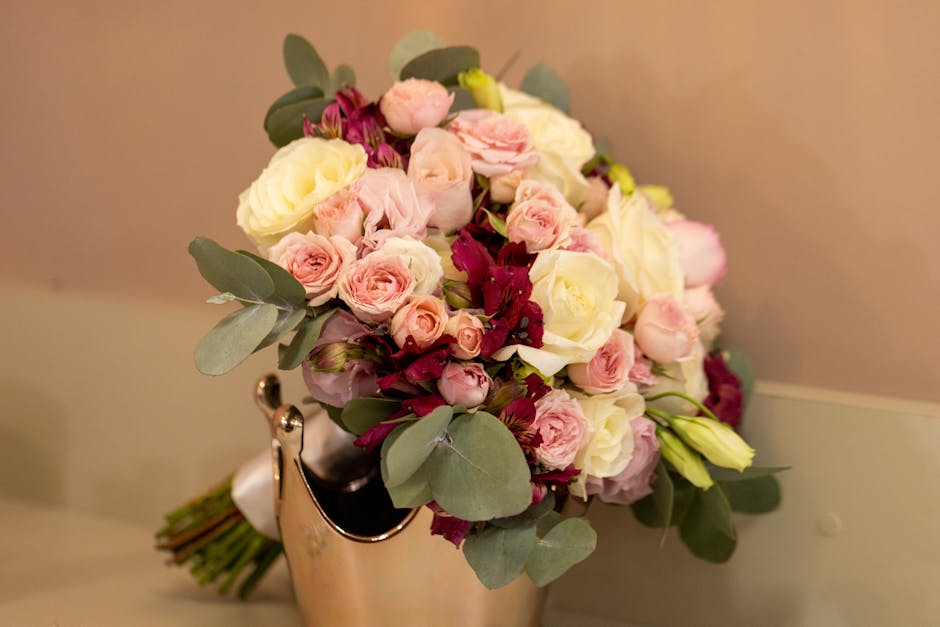 A bouquet arrangement of fresh flowers featuring pale pink roses, cream-colored roses, and deep burgundy blossoms, accented with eucalyptus leaves. The flowers are arranged in a rounded, slightly over