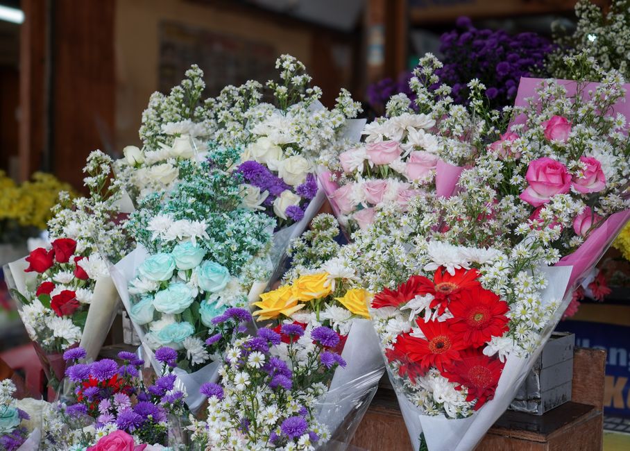 A vibrant display of floral bouquets arranged on a wooden surface inside a flower shop, featuring a variety of fresh flowers including pink roses, red gerberas, white daisies, purple asters, blue rose