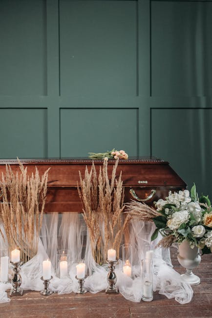 A floral arrangement featuring white roses, peonies, and greenery in a white ceramic vase, positioned on a wooden table alongside tall, wispy pampas grass and lit pillar candles in glass holders. The 